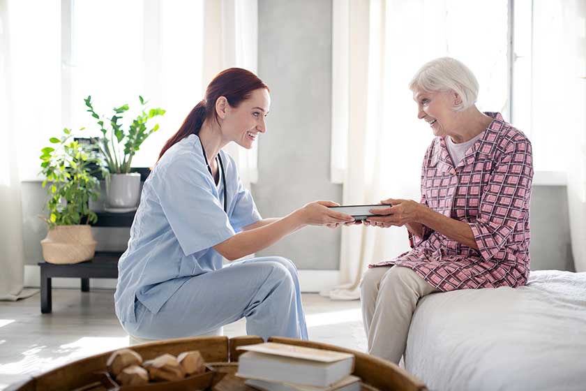 6 Reasons Why 24 Hour Senior Care - Caring Nurse Smiling While Giving Book To Aged Woman